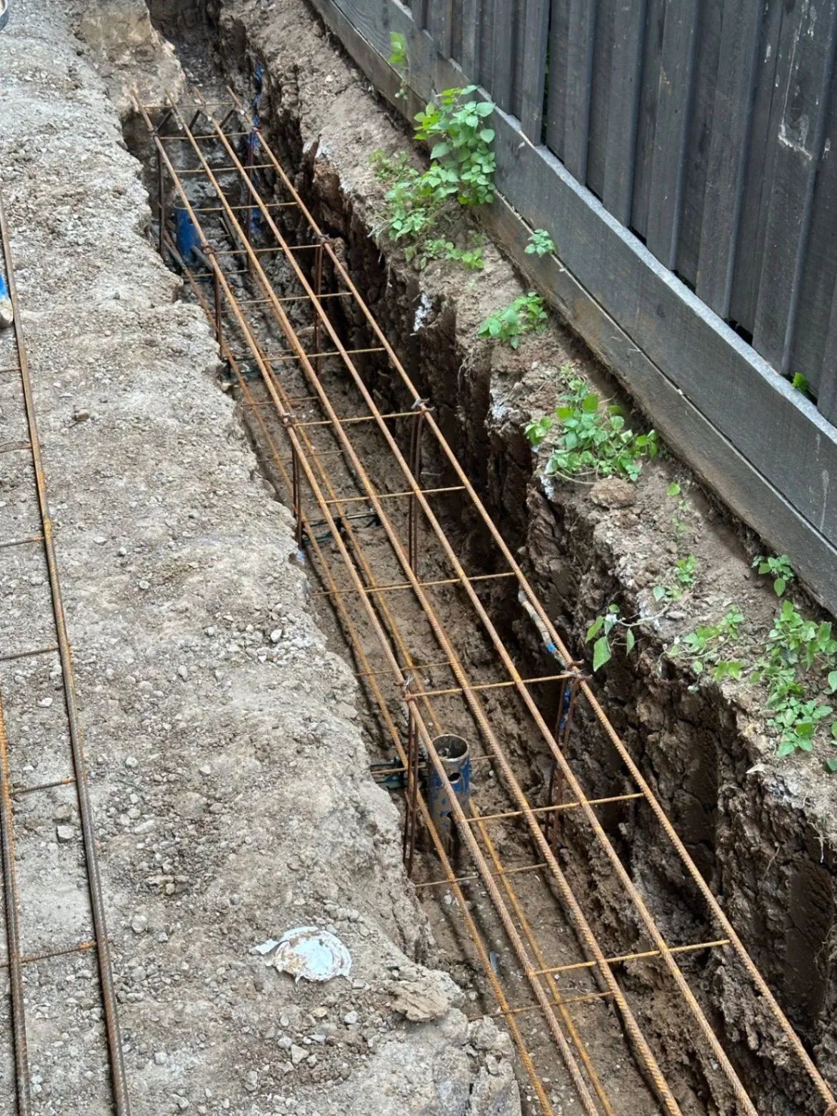 Reinforcing steel set within an excavated trench beside a boundary fence